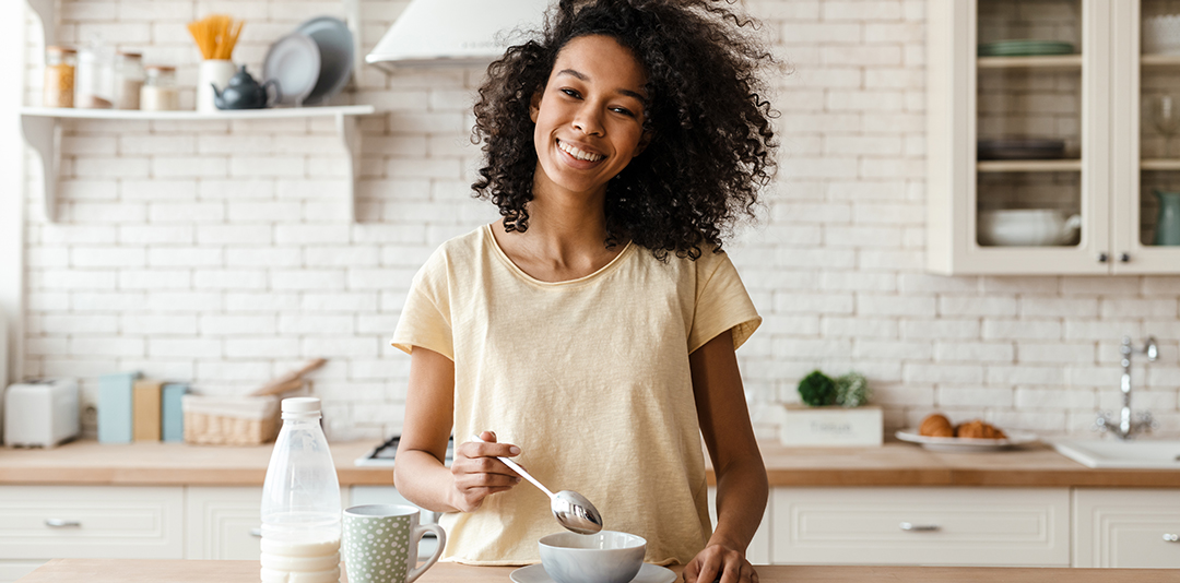Smiling young african woman eating cereal from a bowl while standing at the kitchen counter